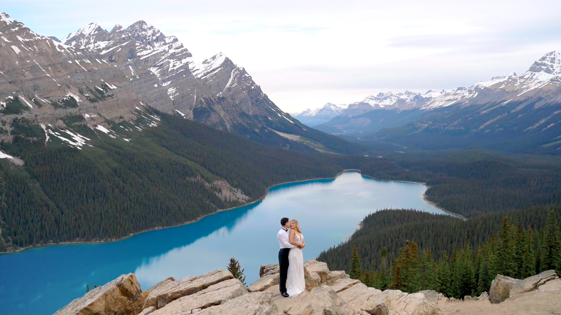 "A couple standing on a rocky outcrop overlooking the turquoise Peyto Lake, surrounded by snow-capped mountains and dense forests.